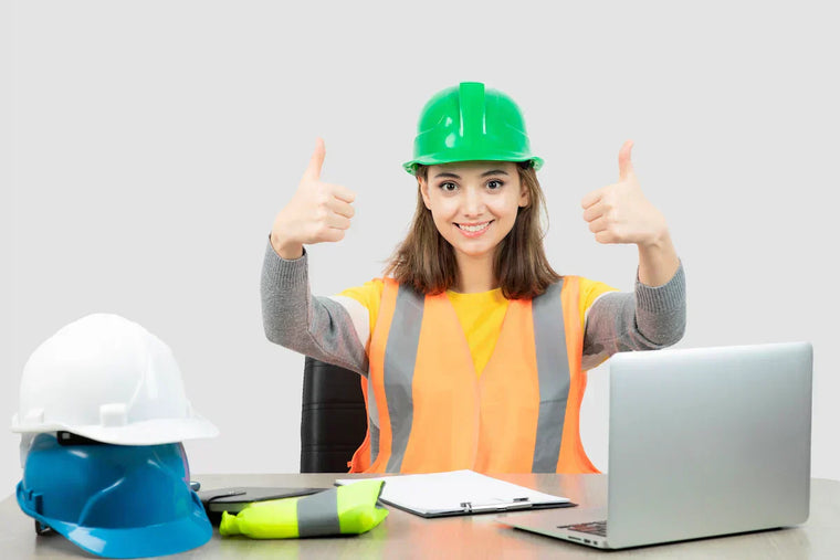 Smiling female safety worker wearing a hard hat and high-visibility vest gives two thumbs up while sitting at a desk with safety equipment.