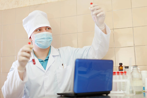 Masked laboratory technician wearing gloves examines test tubes beside a laptop during aseptic laboratory practice.